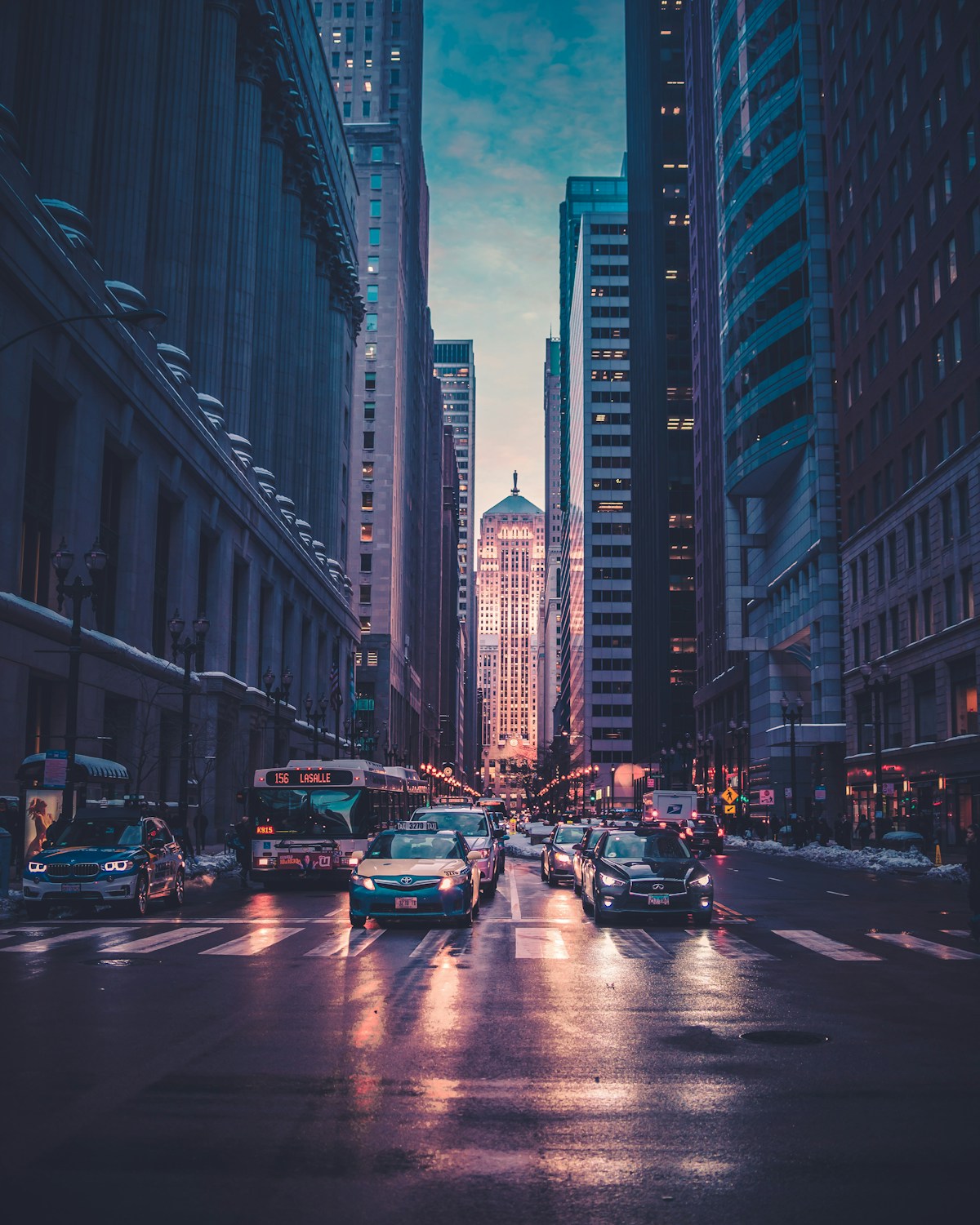 Avenida urbana entre edifícios altos, com veículos e luzes ao entardecer